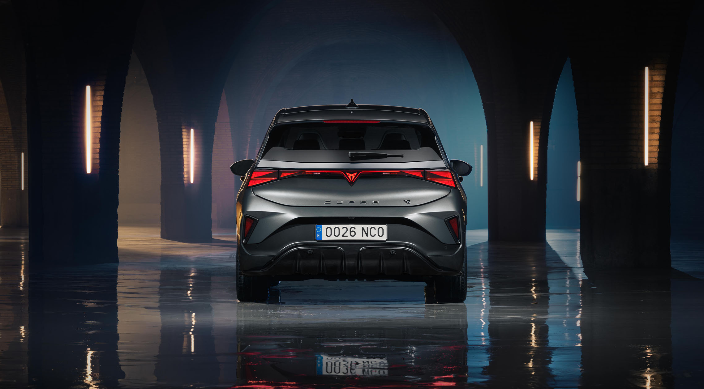 Widok z tyłu samochodu CUPRA Born VZ w kolorze Timanfaya Grey, zaparkowanego w ciemnej, sklepionej przestrzeni przemysłowej z kamiennymi łukami i pionowym oświetleniem neonowym. Zdjęcie podkreśla  listwę tylnych świateł LED, charakterystyczne logo CUPRA oraz odbicie pojazdu na mokrej, wypolerowanej betonowej podłodze. 