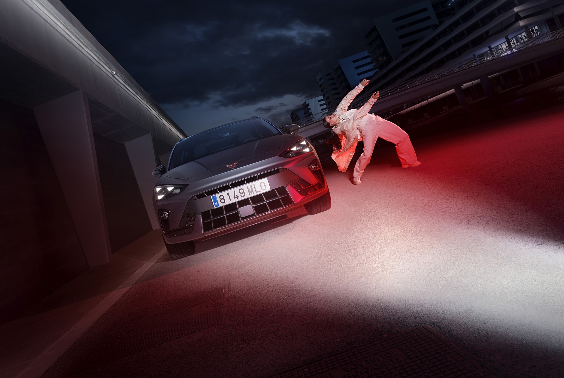 front view of the CUPRA Leon SP in Magnetic Tech Matt Grey colour, riding at night in a city, and an individual next to the vehicle.