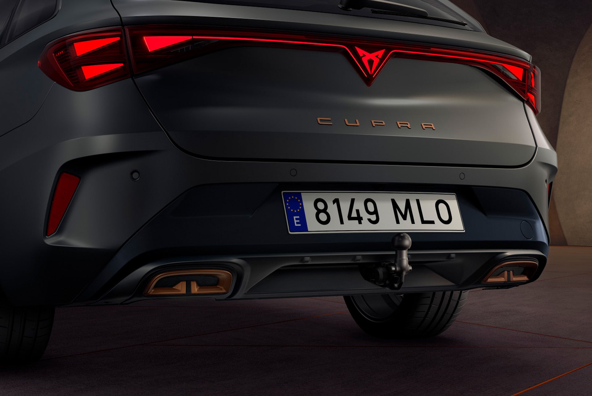 Zbliżenie tylnej części samochodu hybrydowego CUPRA Leon w kolorze Enceladus Grey Matt, z akcentami miedziowymi na felgach, betonową konstrukcją i panoramą miasta w tle