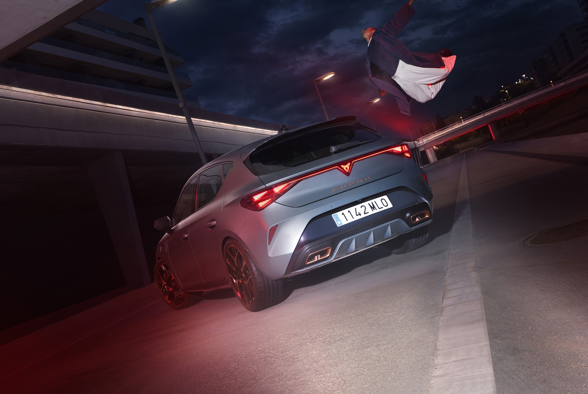 Widok z góry, 3/4 tylnej części samochodu CUPRA Leon PA 5D w kolorze Enceladus Grey Matt. Pojazd jest zaparkowany w miejskiej nocnej scenerii 