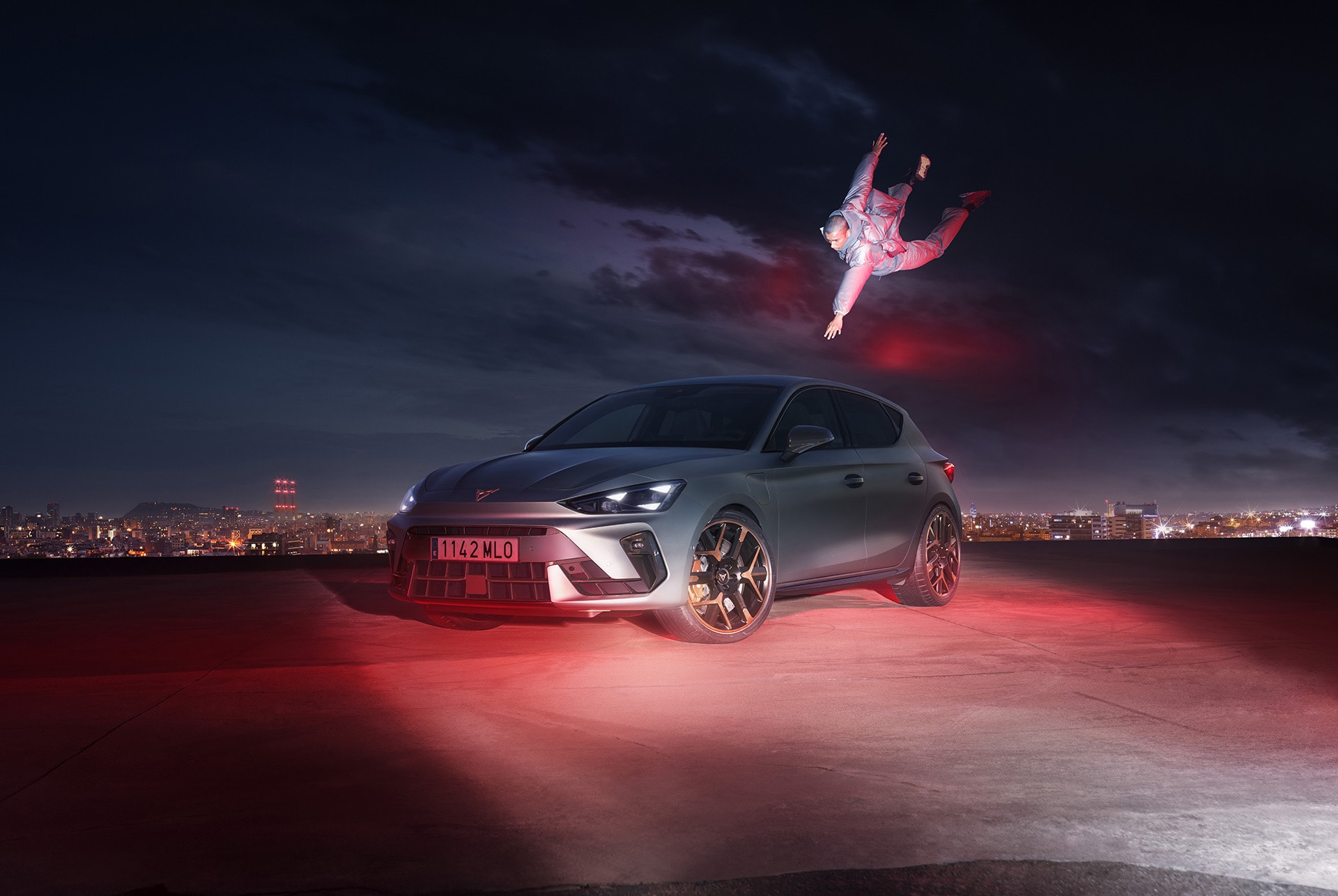 hero angle shot of the CUPRA Leon PA 5D in Enceladus Grey Matt showcasing details such as the 19-inch alloy wheels in Hailstorm Copper tone. The car is shown at night, in a urban setting, and there's an individual near it.