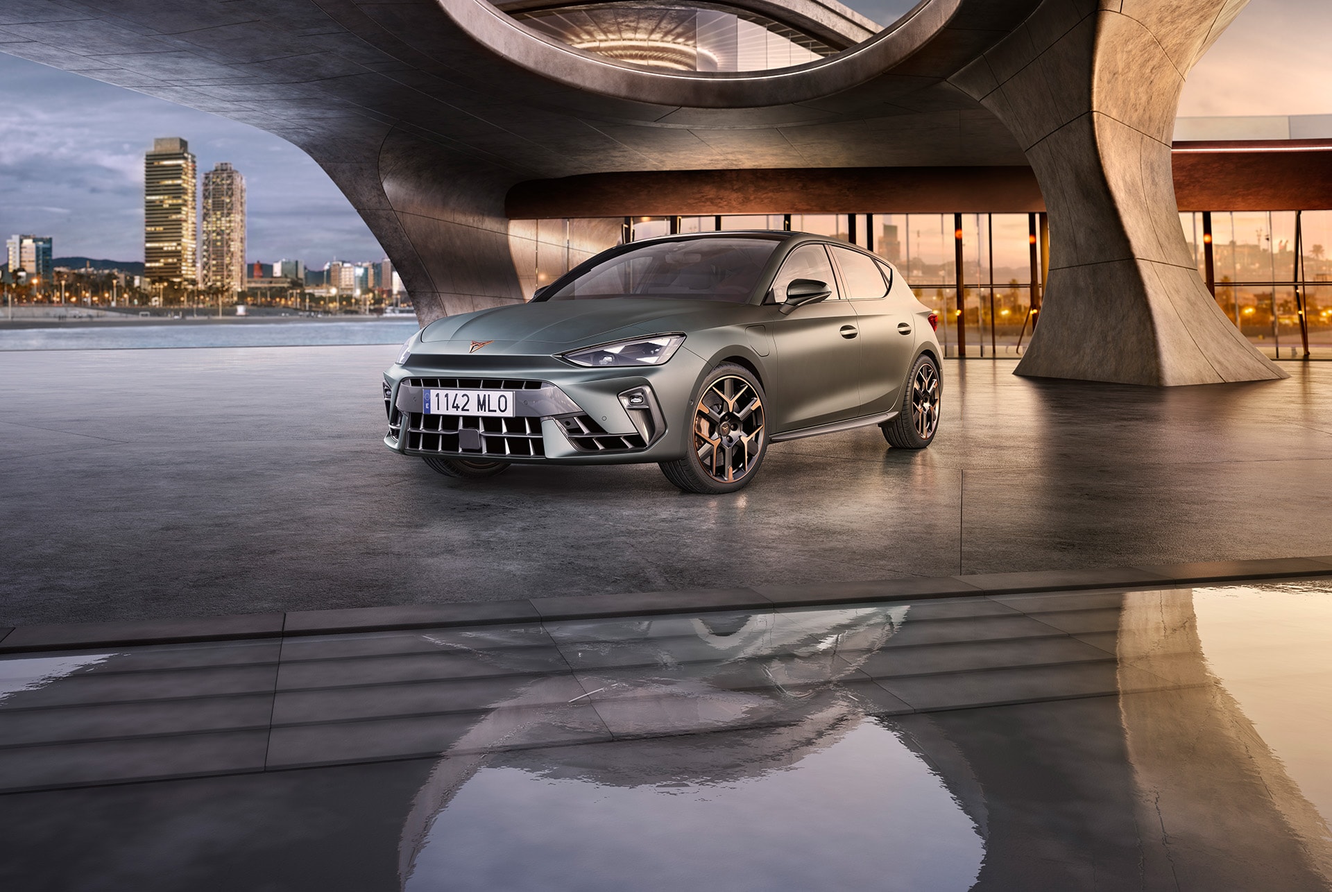 Nowy CUPRA Leon 2024 w kolorze Enceladus Grey Matt z aluminiowymi obręczami kół z miedzianymi akcentami, zaparkowany na gładkim betonie pod konstrukcją architektoniczną. Pejzaż Barcelony w tle.